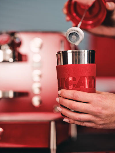 Red Ducati-branded travel mug held below a white nozzle attachment, red Barista M3 blurred behind