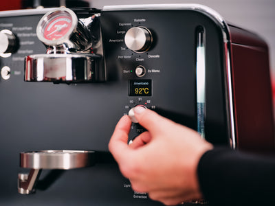 Hand turning the temperature dial on the black control panel, display reading Americano 92°C