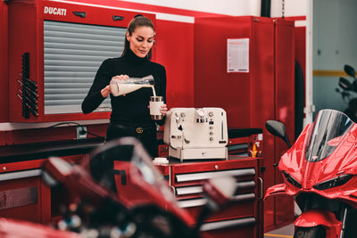 Woman pouring milk into a cup beside the white Barista M3 in a Ducati workshop with a red bike