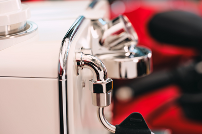 Chrome steam wand and brew head of the white Barista M3 in close-up against a red backdrop