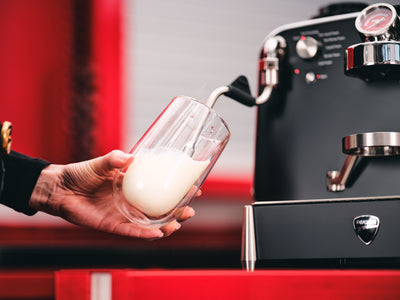 Hand holding a double-walled glass half full of milk beside the black Barista M3 steam wand